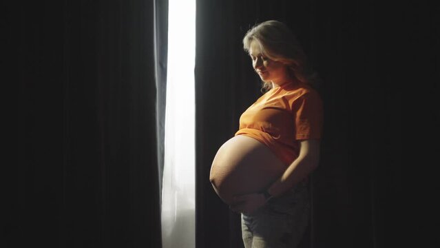 Beautiful Caucasian pregnant woman. The girl strokes her stomach, in which the cavity lives. Difficult last days of pregnancy. A woman strokes her big belly. Happy person. The emergence of a new life
