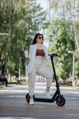 Beautiful young woman in white suit and sunglasses standing with her electric scooter in city parkland and drinking coffee