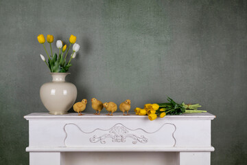 Spring bouquet of white and yellow tulips in a vase with little baby chickens on vintage green wall background - easter still life with white carved wooden fireplace