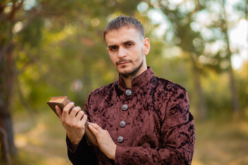 A young man in medieval brown clothes stands with an old book in his hand. A guy in a historical costume in nature.