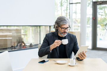 Bearded mature economist or banker in suit and eyeglasses analyzing online financial data in tablet by cup of coffee in modern restaurant