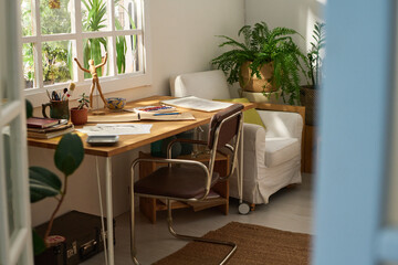 Part of room or home studio of modern artist with sketches on papers, notepads and other creative items on table standing by window