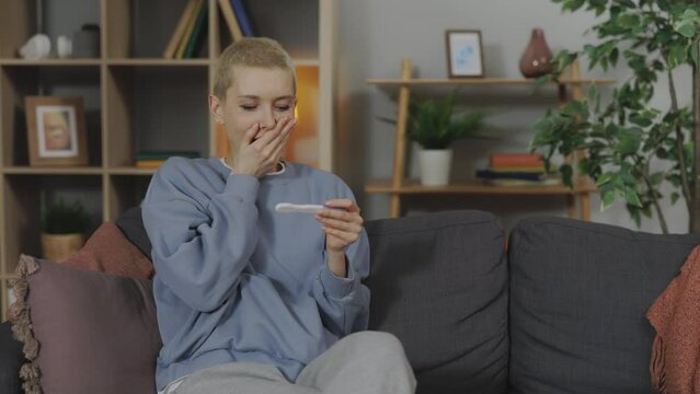 Surprised caucasian woman checking pregnancy test while sitting on couch. Beautiful lady with short blond hair feeling happiness during long-awaited pregnancy.