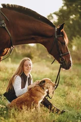 Deurstickers A young beautiful woman jockey with her dog sits in a meadow near her horse at sunset. © sergo321