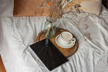 Cozy breakfast in bed. Cup of hot cappuccino on wooden tray, bouquet of dried flowers in glass vase, book, tablet. Work online at home.