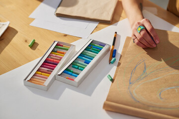 Close-up of hand of young creative female artist sketching on paper or notepad with blue crayon while sitting by wooden table in studio