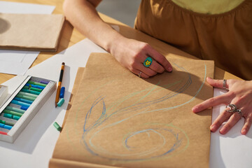 Close-up of young creative woman drawing sketch on paper with blue crayon while sitting by...