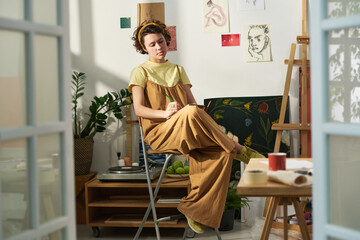 Young inspired female artist making sketches of painting subjects in notepad while sitting against...