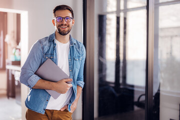 Portrait of a young modern businessman standing holding a laptop and looking at the camera with a...