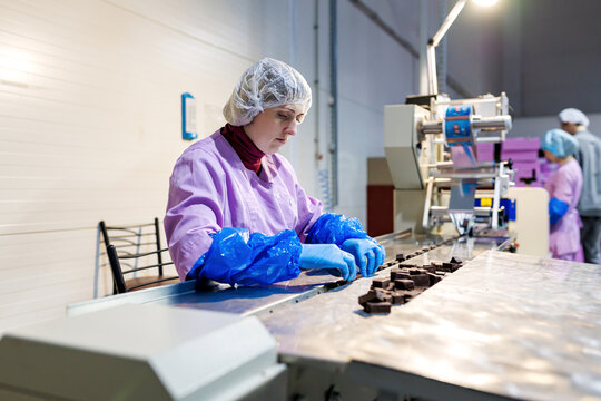 Professional female worker in uniform and protective gloves sorts chocolate candies on production line at factory