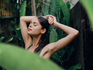 Fototapeta na wymiar A woman takes a shower and washes her head and hair outdoors in nature, closed eyes and a smile on the background of tropical plants, palm trees, green banana leaves, summer rain, lifestyle