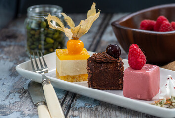 set of cakes, tangerine, chocolate and raspberry flavors, on a white rectangular plate, worn wooden background in blue tones.