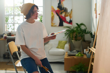 Young serious woman with color palette looking at panel for painting standing by wall while thinking of new creative ideas in studio