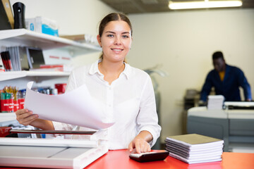 Cheerful young woman in white shirt smiling at the camera and using paper cutter in the printing...