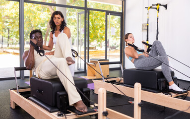 Focused Latina instructor helping African American man practicing pilates on reformer during group classes at fitness studio