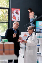 Elderly pharmacist helping client with pharmaceutical products trying to find cardiology drugs in pharmacy. Customer looking at medicaments on drugstore shelves. Medicine service and concept