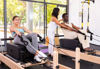 Positive woman and African American man training stretching exercises on reformer with female instructor at gym