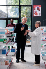 Senior specialist explaining cardiology pills treatment to old man customer, holding box of medicine and read medical leaflet. Client asking about pharmaceutics products in pharmacy shop.