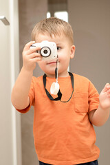 Fototapeta premium People. Handsome funny and emotional caucasian blond little boy in an orange t-shirt with a children's white camera takes pictures in the home interior.