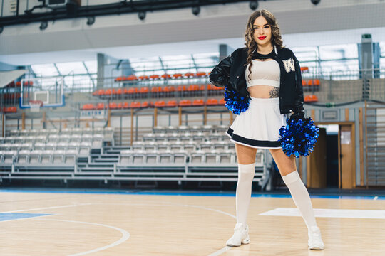 Full shot of a cheerleader in uniform with a jacket posing on basketball court. Sports hall's boxes blurred in the background. High quality photo