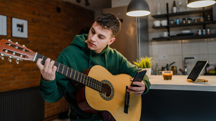One young man Caucasian teenager sit at home in room playing guitar use smartphone mobile phone to...
