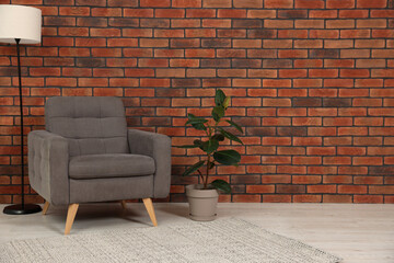 Cosy armchair, lamp and potted plant near brick wall in room, space for text. Interior design