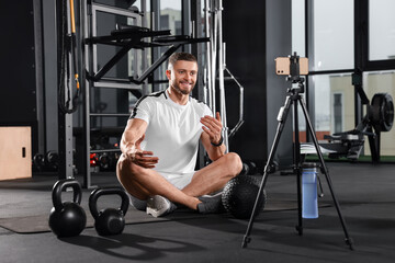 Fototapeta premium Man with ball and dumbbells streaming online training on phone at gym. Fitness coach