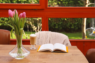 Glass of delicious cocoa, pink tulips and book on wooden table at terrace