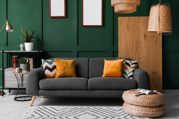 Interior of living room with black sofa, wicker pouf and blank frames near green wall