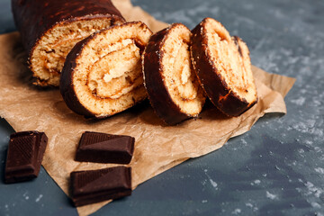 Baking paper with pieces of delicious sponge cake roll and chocolate on grey table