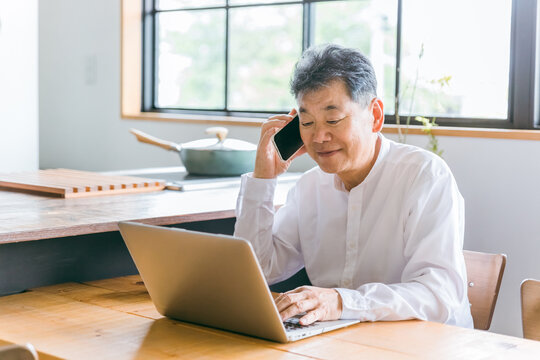 家のダイニングでノートパソコンとスマホ・スマートフォンを使って電話する高齢者男性（アジア人・中国人）