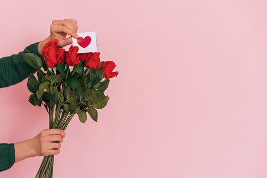 Female hand with bouquet of red roses against pink background.