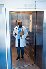 Fototapeta premium Vertical of african american male doctor using smartphone in hospital lift with copy space