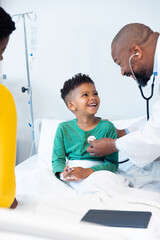 African american male doctor using stethoscope on boy patient in hospital with copy space