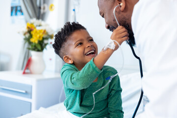 Laughing african american boy patient taking male doctor's stethoscope in hospital - Powered by Adobe
