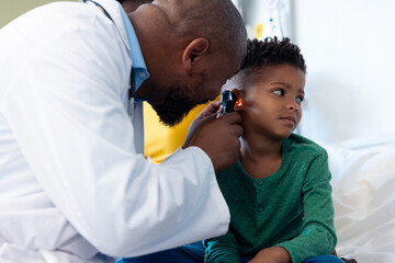 African american male doctor using otoscope to examine ear of boy patient in hospital - obrazy, fototapety, plakaty