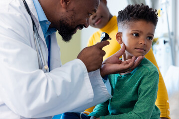 African american male doctor using otoscope to examine ear of boy patient, with mother