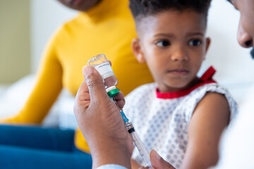 African american male doctor preparing vaccine for boy patient, with mother in hospital