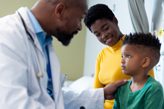 African american male doctor talking to boy patient, with mother in hospital
