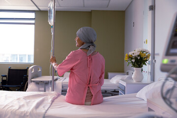 Senior caucasian female cancer patient wearing head scarf sitting on bed with drip at hospital