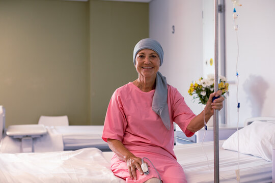 Happy senior caucasian female cancer patient wearing head scarf sitting on bed at hospital