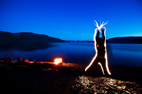 A woman sends energy through her body and out her hands at sunset next to a campfire and lake in Idaho. This light painting image was created with a long exposure and flash lights.