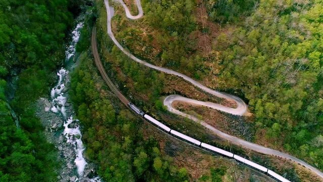 Train rolling down the railroad tracks in a forest between a river and a winding road - aerial flyover