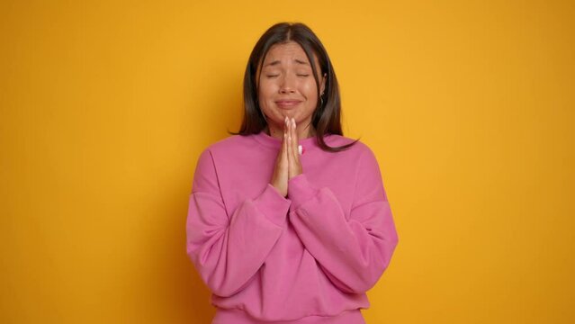 Asian young woman doing praying gesture with hands and begging in front of camera, showing desire and expectation sign. imploring and asking with palms and body language , isolated next to yellow