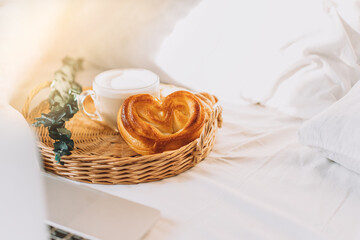 Wicker tray with croissants and coffee on white bed linen with laptop.