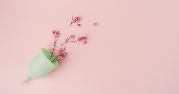Top view dry red flowers in green menstrual cup against pink background.