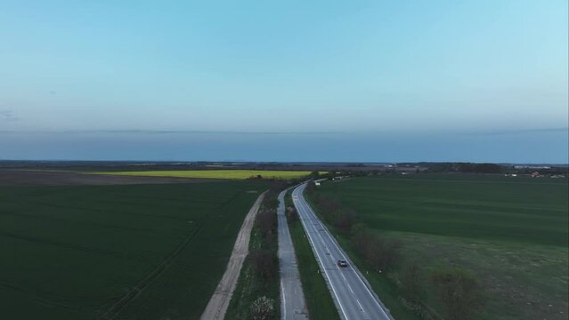 Aerial cinematic view of cars driving between the trees and fields on countryside roadway with rural roads on the side.Drone follow fottage during twilight in summer.Lights on the cars.Coming home