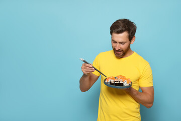 Happy man holding plate with tasty sushi rolls and chopsticks on light blue background. Space for text