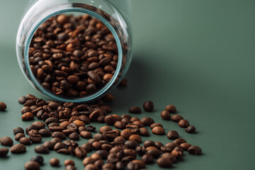 Coffee beans pour out of a glass jar on a green background.