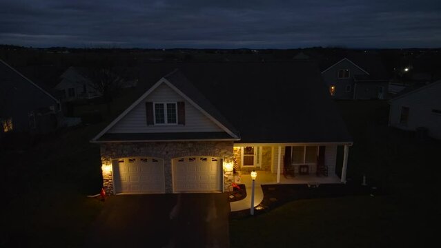 Lights on exterior of house lit up during blue hour in American neighborhood. Dark, aerial rising shot at night of home.